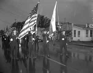 Flags wave, guys march down wet street.