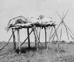 Three wrapped bodies on top of raised scaffolding with tipi frame in background.